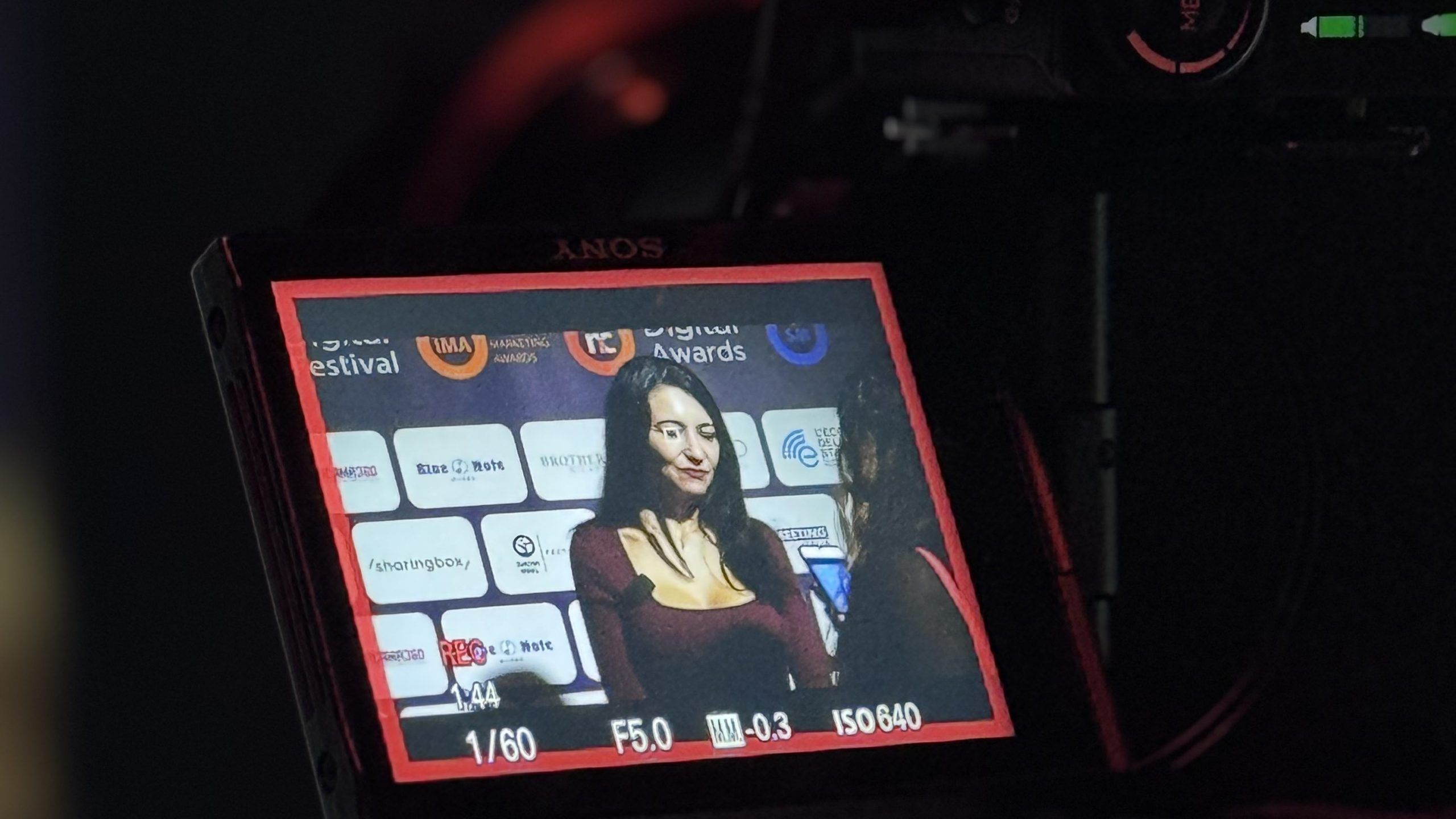 Interview at a digital awards event shown through the display of a Sony camera. The background features various sponsor and organizer logos. A woman in a formal outfit is being interviewed by a journalist. Interview at a digital awards event shown through the display of a Sony camera. The background features various sponsor and organizer logos. A woman in a formal outfit is being interviewed by a journalist.